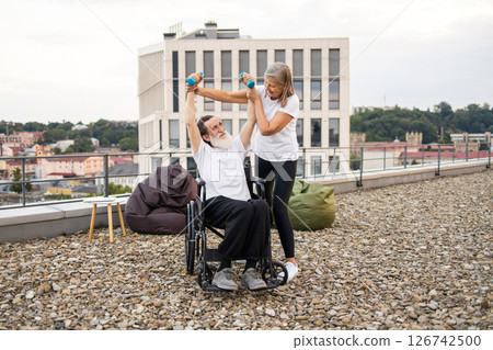 Senior woman assisting wheelchair-bound husband with rehabilitation exercises using dumbbells on rooftop. Bright day enhancing supportive, emotional interaction, emphasizing recovery, togetherness. Senior woman assisting wheelchair-bound husband with rehabilitation exercises using dumbbells on rooftop. Bright day enhancing supportive, emotional interaction, emphasizing recovery, togetherness. 126742500