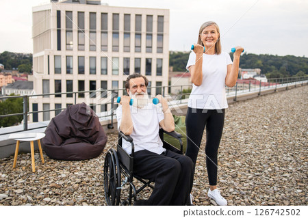 Older couple engaging in light exercise with dumbbells, promoting recovery and health. Woman demonstrates activity while helping seated husband in wheelchair. Emphasizes fitness, rehabilitation Older couple engaging in light exercise with dumbbells, promoting recovery and health. Woman demonstrates activity while helping seated husband in wheelchair. Emphasizes fitness, rehabilitation 126742502