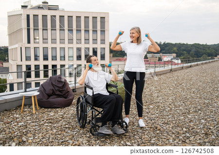 Senior man in wheelchair exercising with dumbbells assisted by woman on building rooftop. They are enjoying fitness outdoors together, focusing on health and rehabilitation in inclusive environment. 126742508
