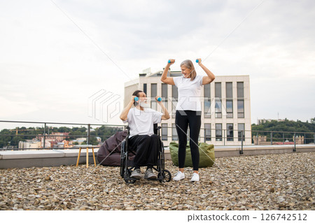Senior man in wheelchair and woman in casual attire perform exercises with dumbbells outdoors in urban setting on gravel rooftop. Concept of health, fitness outdoor activity, and rehabilitation. 126742512