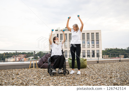 Senior man in wheelchair and woman exercising with dumbbells outdoors on urban rooftop promoting active lifestyles. Encouragement, togetherness, health, vitality, and support in fitness 126742514