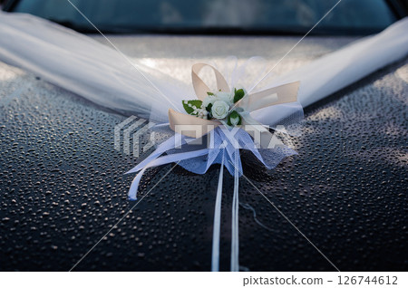 Elegant wedding car decoration featuring white tulle draping over the hood with a delicate floral arrangement of roses and ribbons, wet surface reflecting light Elegant wedding car decoration featuring white tulle draping over the hood with a delicate floral arrangement of roses and ribbons, wet surface reflecting light 126744612