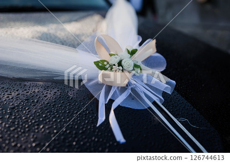 Wedding car decoration with a delicate white tulle bow and floral arrangement featuring roses, ribbons, and a heart-shaped detail black car roof 126744613