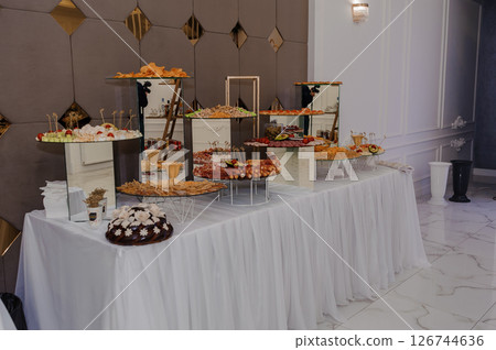 Elegant buffet table arrangement in a luxurious hall with white linens, tiered stands displaying various appetizers and desserts, ornate decor and a polished marble floor setting Elegant buffet table arrangement in a luxurious hall with white linens, tiered stands displaying various appetizers and desserts, ornate decor and a polished marble floor setting 126744636