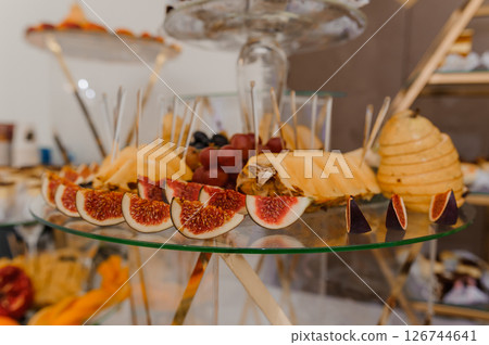 Elegant fruit and cheese platter presentation on a golden stand, featuring figs, grapes, pears, and a honeycomb, styled for a luxurious event or catered gathering Elegant fruit and cheese platter presentation on a golden stand, featuring figs, grapes, pears, and a honeycomb, styled for a luxurious event or catered gathering 126744641