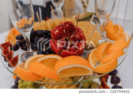 Elegant fruit platter arrangement with oranges, pomegranates, grapes, figs, and wine glasses on a white table setting, creating a festive and luxurious atmosphere 126744642