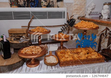 A lavish snack buffet is displayed on a table with wooden stands holding various chips and savory bites, decorated with nautical elements like a blue crab and fishing tools 126744647