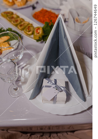 Elegant wedding table setting with a folded napkin, small gift box, glassware, and appetizers on a white tablecloth, creating a festive and sophisticated atmosphere 126744649