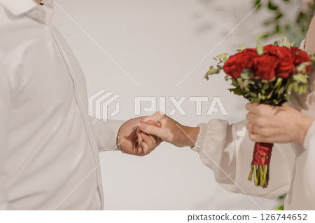 A tender moment capturing a wedding ceremony with two people holding hands, symbolizing love and commitment, while a bouquet of vibrant red roses adds a touch of romance to the scene, interior setting 126744652