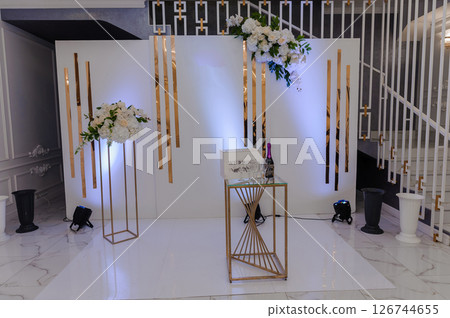 Elegant Wedding Backdrop Setup with White Floral Arrangements and Gold Accents in a Modern Event Space, featuring a Bottle of Champagne and Decorative Elements Elegant Wedding Backdrop Setup with White Floral Arrangements and Gold Accents in a Modern Event Space, featuring a Bottle of Champagne and Decorative Elements 126744655