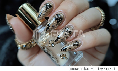 close-up of a woman's hand holding a bottle of nail polish. The nails are painted with a gold glittery polish and have a unique design. The top of the nails are decorated with small black	 126744711