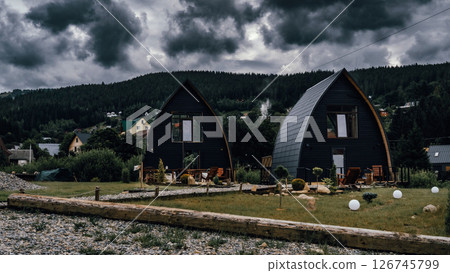 Two modern A-frame cabins sit side by side under a dramatic, stormy sky. Two modern A-frame cabins sit side by side under a dramatic, stormy sky. 126745799