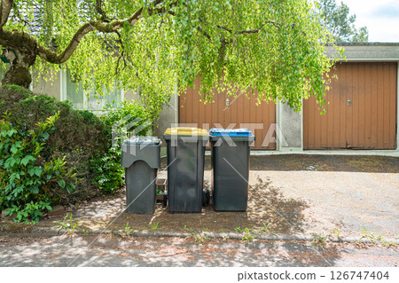 Three recycling bins with different colored lids stand under a lush tree in front of closed garage doors. The setting highlights the importance of waste sorting in residential areas. Three recycling bins with different colored lids stand under a lush tree in front of closed garage doors. The setting highlights the importance of waste sorting in residential areas. 126747404