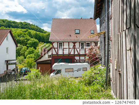 Charming half-timbered house with a camper van parked nearby, set in a lush green landscape on a sunny day. Traditional architecture meets modern travel in a peaceful countryside setting. Charming half-timbered house with a camper van parked nearby, set in a lush green landscape on a sunny day. Traditional architecture meets modern travel in a peaceful countryside setting. 126747405
