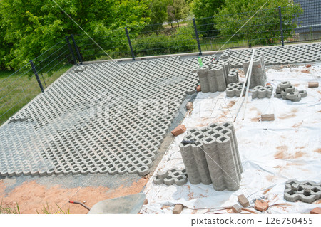Concrete grid blocks are laid out at a construction site for a new driveway. Stacks of blocks and tools are visible, indicating ongoing work. Trees provide a lush background. 126750455