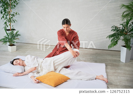 Masseuse making thai yoga massage. Woman therapist doing traditional massage treatment, stretching female patient's body. Client lying on mat, enjoying hand massage in calm atmosphere. 126752315