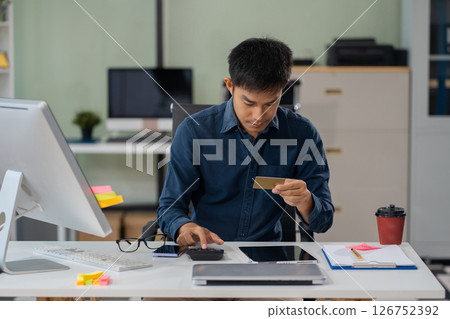 Asian man stressed over credit card debt, holding bills in frustration. Financial anxiety and economic burden concept in a realistic lifestyle setting. 126752392