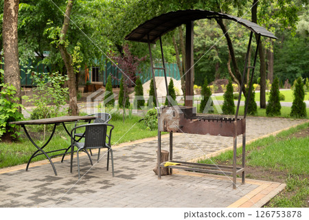 Outdoor barbecue grill with canopy, metal table, and chairs in a green garden setting, perfect for summer picnics and gatherings. 126753878