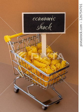 ECONOMIC SHOCK text on Blackboard label against Shopping trolley cart Filled With Pasta on Beige background. Food and groceries shopping price increase, Rising food cost food crisis inflation ECONOMIC SHOCK text on Blackboard label against Shopping trolley cart Filled With Pasta on Beige background. Food and groceries shopping price increase, Rising food cost food crisis inflation 126754701