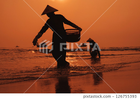Silhouette of a Vietnamese woman in straw hat on shore of sea with ships in fishing village at sunset in Vietnam 126755393