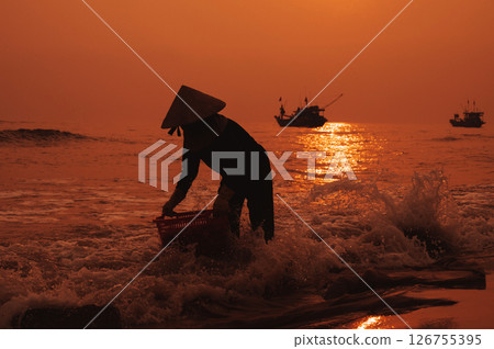 Silhouette of a Vietnamese woman in a straw hat on the shore of sea with ships in a fishing village in morning at sunrise in Vietnam 126755395