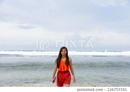 Confident young asian woman strides along beach near huge sea waves 126755581