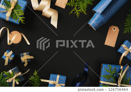Frame of blue gifts with gold ribbons, brown paper tags, green thuja branches on black background. Space for your text. 126756464