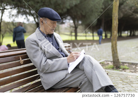 Elderly writer taking notes while listening to music on a park bench Elderly writer taking notes while listening to music on a park bench 126757730