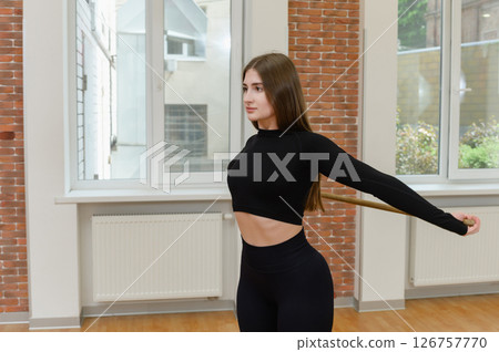 Young woman stretching shoulders with wooden stick indoors. 126757770