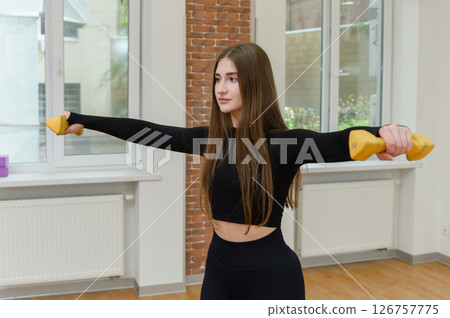 Woman doing lateral raise with yellow dumbbells indoors. 126757775
