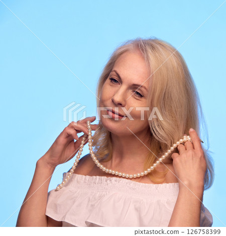 Smiling senior woman gracefully holds pearl necklace near neck, enjoying soft touch and confident elegance on blue background Smiling senior woman gracefully holds pearl necklace near neck, enjoying soft touch and confident elegance on blue background 126758399