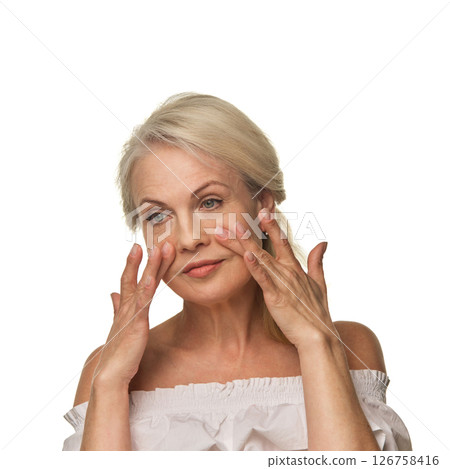 Elderly woman uses fingertips to massage skin under eyes against white background, reducing puffiness and supporting skincare routine with confidence 126758416