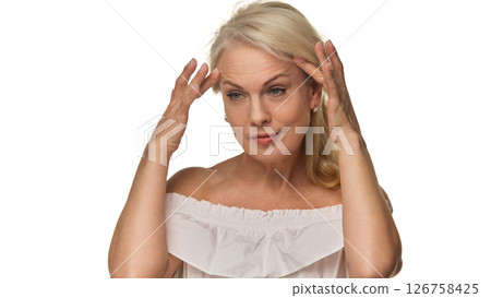 Senior woman gently touches temples while evaluating skin with focused expression against white background, showing beauty and self-acceptance 126758425
