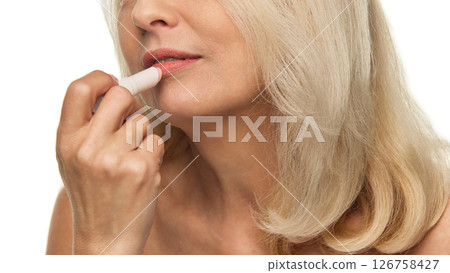 Elderly woman softly applies lip balm to lips with gentle motion, showing hydrated skin and soft glow against white studio background 126758427