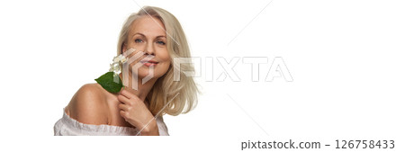 Mature woman holding flower near face with relaxed look showing radiant skin against white studio background with negative space. Banner Mature woman holding flower near face with relaxed look showing radiant skin against white studio background with negative space. Banner 126758433