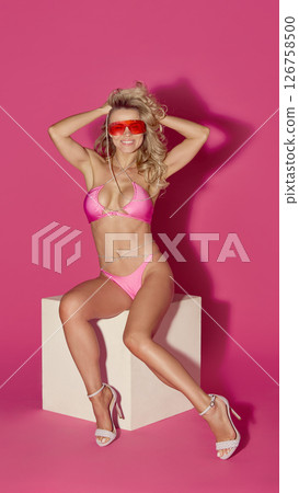 Smiling woman in pink bikini lifts hands behind head while sitting on cube in red sunglasses against pink background 126758500