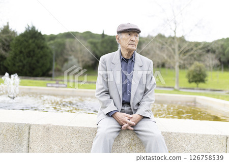 Elderly man sitting pensively by a fountain in a park Elderly man sitting pensively by a fountain in a park 126758539
