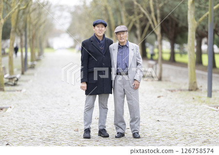 Adult son standing next to elderly father in park during winter Adult son standing next to elderly father in park during winter 126758574