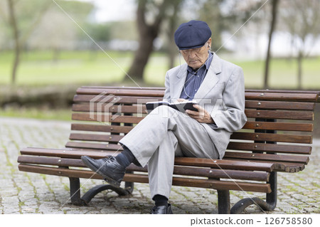 Elderly writer taking notes in a notebook while sitting on a bench in the park Elderly writer taking notes in a notebook while sitting on a bench in the park 126758580