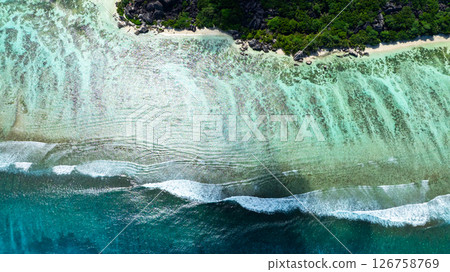 Waves reach the clear sandy shoreline bordered by lush green vegetation from an aerial perspective. Seychelles, La Digue. Waves reach the clear sandy shoreline bordered by lush green vegetation from an aerial perspective. Seychelles, La Digue. 126758769