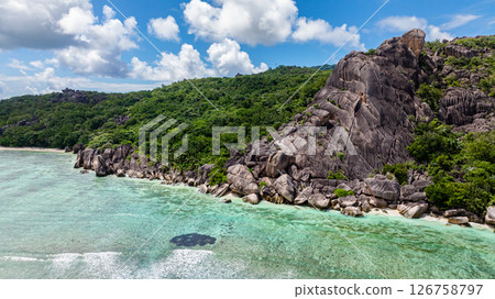 Steep cliffside covered with lush green foliage, overlooking turquoise waters with gentle waves under a clear sky. Seychelles, La Digue. Steep cliffside covered with lush green foliage, overlooking turquoise waters with gentle waves under a clear sky. Seychelles, La Digue. 126758797
