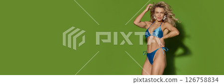 Smiling blonde woman, wearing in blue bikini posing with hand on hip against green background. Banner with copy space 126758834