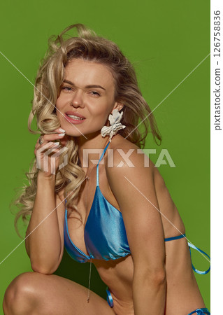 Close-up portrait of sensual blonde woman in shiny blue bikini looking at camera with confidence against green studio background. Close-up portrait of sensual blonde woman in shiny blue bikini looking at camera with confidence against green studio background. 126758836