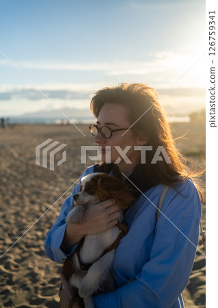 A young woman hugs her Cavalier King Charles Spaniel on the beach at sunset. Warm light, waves, and smiles capture a peaceful, joyful moment of connection and relaxation. A young woman hugs her Cavalier King Charles Spaniel on the beach at sunset. Warm light, waves, and smiles capture a peaceful, joyful moment of connection and relaxation. 126759341