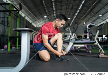 Wellness and Fitness. Man tying shoelaces in gym, preparing for workout. Wellness and Fitness. Man tying shoelaces in gym, preparing for workout. 126759864