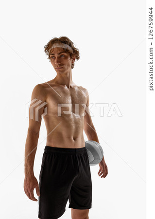 Shirtless male athlete with confident look holds volleyball at waist while standing tall in black shorts against white studio background. 126759944