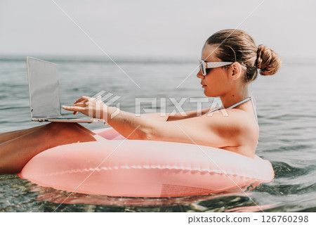 A woman is sitting on a pink inflatable raft in the water, using a laptop. Concept of relaxation and leisure, as the woman is enjoying her time by the water while working on her laptop. 126760298