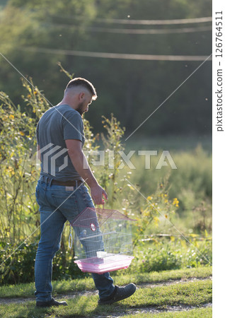 Man carrying a birdcage outdoors on a sunny day. 126764511