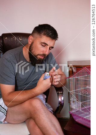Man gently holds a small blue bird. A birdcage sits nearby. 126764513