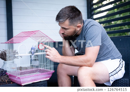 Man gently checks on his pet bird in its cage outdoors. Man gently checks on his pet bird in its cage outdoors. 126764515